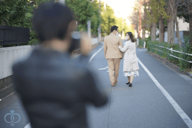 道を歩く男女を背後からカメラで撮影している調査員の画像。浮気調査や婚約者の行動確認といった尾行調査の実施場面を表している。