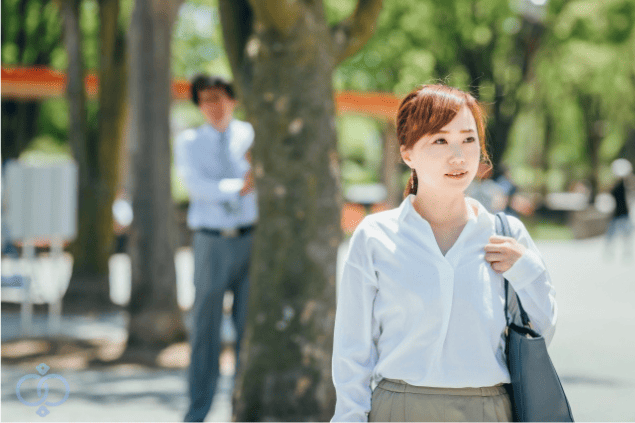 女性の背後から木陰に立つ男性が見つめる様子。隣人によるつきまとい行為が始まる場面の不安感を表現したイメージ。