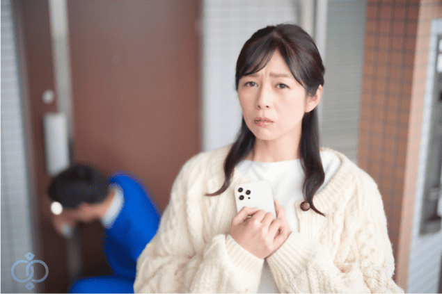 不安げな表情でスマートフォンを手にする女性と、その後ろで玄関付近にいる人物。ストーカー被害の不安や警戒心を表現した場面。