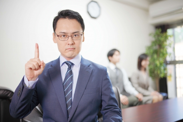 スーツ姿の男性が指を立てて説明している様子。脅迫状への対応事例をもとに注意点や学びを伝えているイメージ。