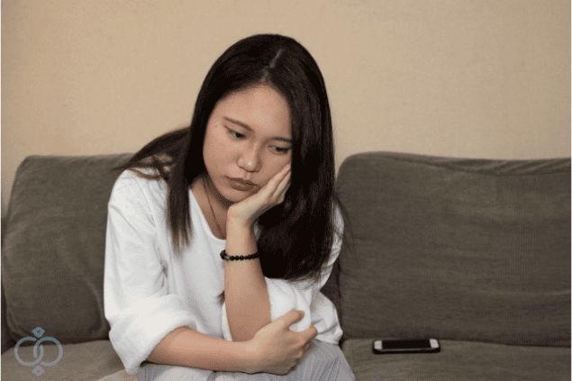 ソファに座ってうつむく女性と横に置かれたスマホ。彼氏が泊まりに来なくなった違和感から身辺調査を依頼するきっかけを表現。