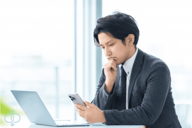 スマートフォンを見つめて考え込むスーツ姿の男性。貸したお金が返ってこないことに悩んでいる状況をイメージした写真。