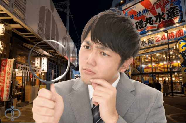 繁華街の背景でルーペを持ち、真剣な表情で調べている男性の画像。風俗嬢にお金を貸した状況で、本当か利用されているかを相手の行動で確かめようとしている場面を表現。
