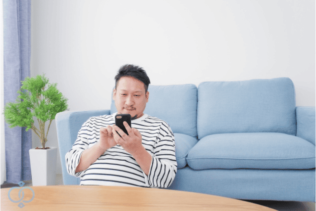 自宅のソファでスマホを見ながら思い出に浸る男性の画像。好きだった風俗嬢を思い出し、なぜ探したいのかを考えている様子を表現。