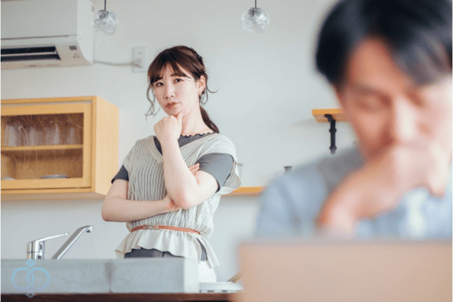 パソコンを見ている男性を後ろから疑わしげに見つめる女性の画像。彼氏の行動に違和感を覚え、既婚者かもしれないと疑い始めた場面を表現している。
