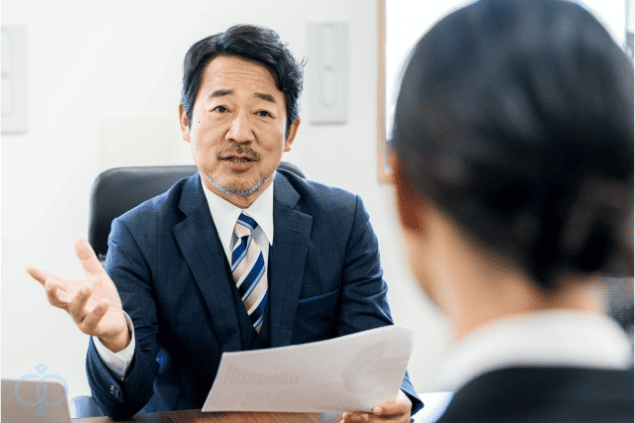 スーツ姿の相談員が資料を手に持ち、依頼者に優しく説明している様子。夫の素行調査相談窓口を示すイメージ画像。