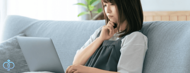 ソファーでパソコンを見ながら考え事をする女性。お金を渡す以外の支援を考える様子をイメージした画像