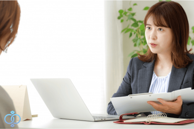 女性スタッフが資料を見せながら説明し素行調査を検討する依頼者に探偵が相談対応している様子のイメージ画像