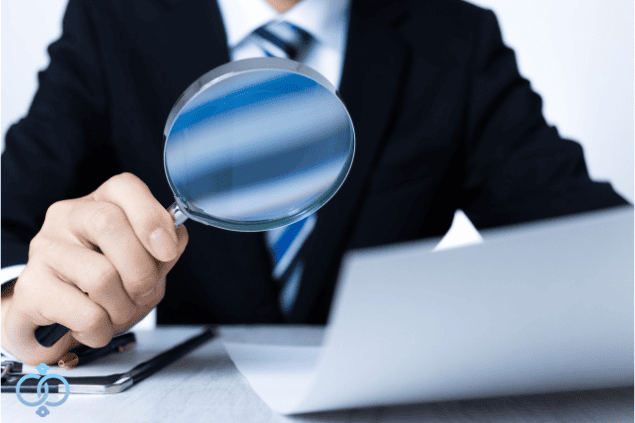 企業調査の基本について、依頼前に知っておきたい情報をパソコンと虫眼鏡で確認するビジネス担当者の様子。