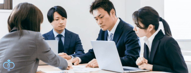 企業調査が必要になるケースについて、書類やパソコンを確認しながらリスクや相談内容を整理するビジネス担当者たちの様子。