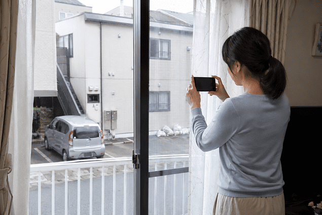 近所の嫌がらせに備えて自宅の窓から屋外の状況を撮影する女性